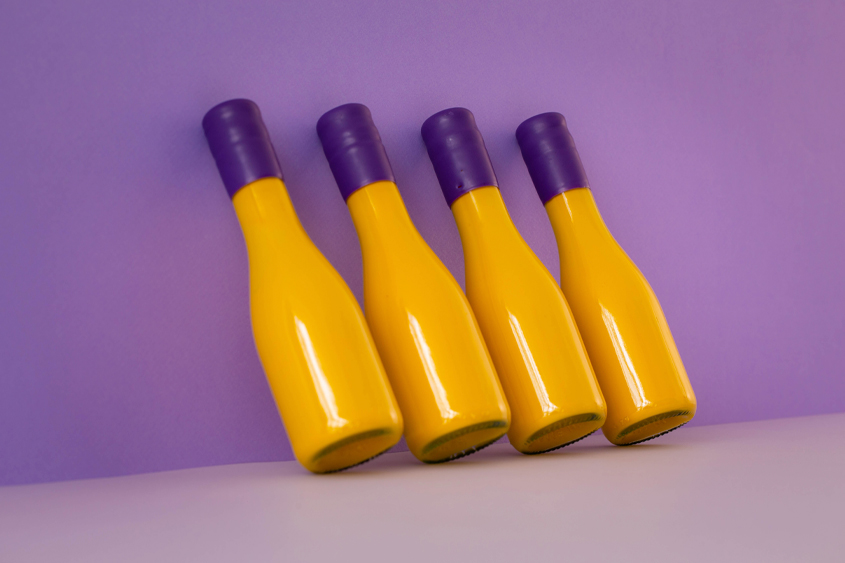 4 botellas inclinadas de color amarillo con lacre color morado en el cuello de la botella y un fondo color morado obscuro y piso color morado claro