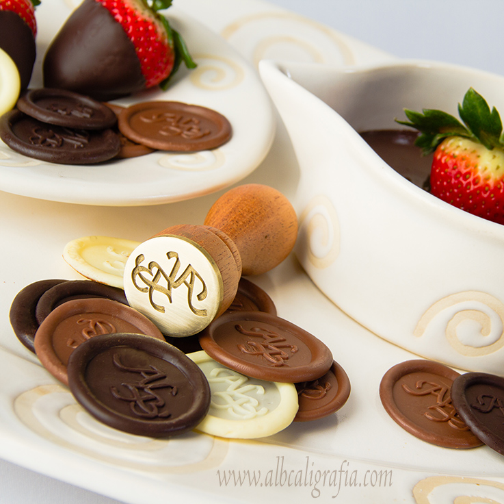 Sello metálico para sellar chocolates, rodeado de chocolates estampados con el sello de ALB y algunas fresas cubiertas con chocolate sobre cerámica bl