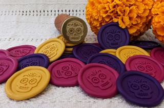 Medallones de lacre de colores magenta, morado y amarillo con el diseño de catrina, acompañadas por el sello para lacrar y unas flores de cempasúchil