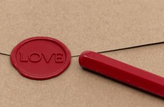 Sobre cafe lacrado con sello de diseño LOVE en color rojo, junto con una barra de lacre para papel un poco derretida de la punta